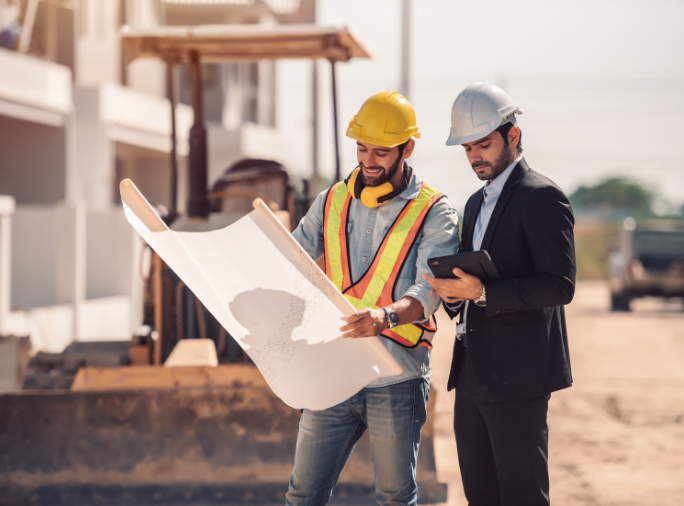 Two professionals reviewing construction plans on a job site, one in safety gear and hard hat, the other in business attire with a tablet.
