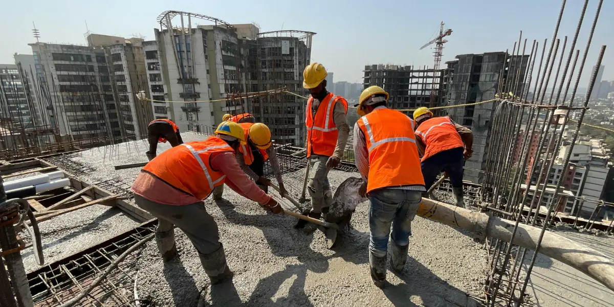 Construction team in safety gear working on a concrete deck at a high-rise construction site with city buildings and crane in background.
